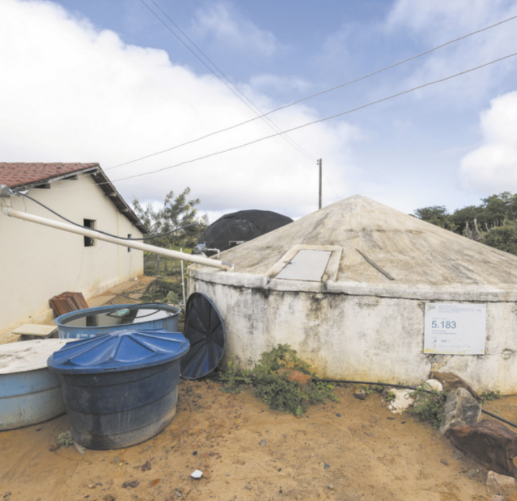 Fotografia de uma grande cisterna com um cano conectado. Ao lado, há uma casa e duas caixas de água.