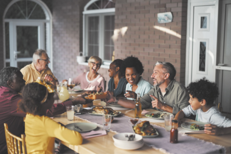 Fotografia de uma família sentada à mesa para fazer uma refeição. A mesa está posta e as pessoas estão sorrindo e conversando entre si.