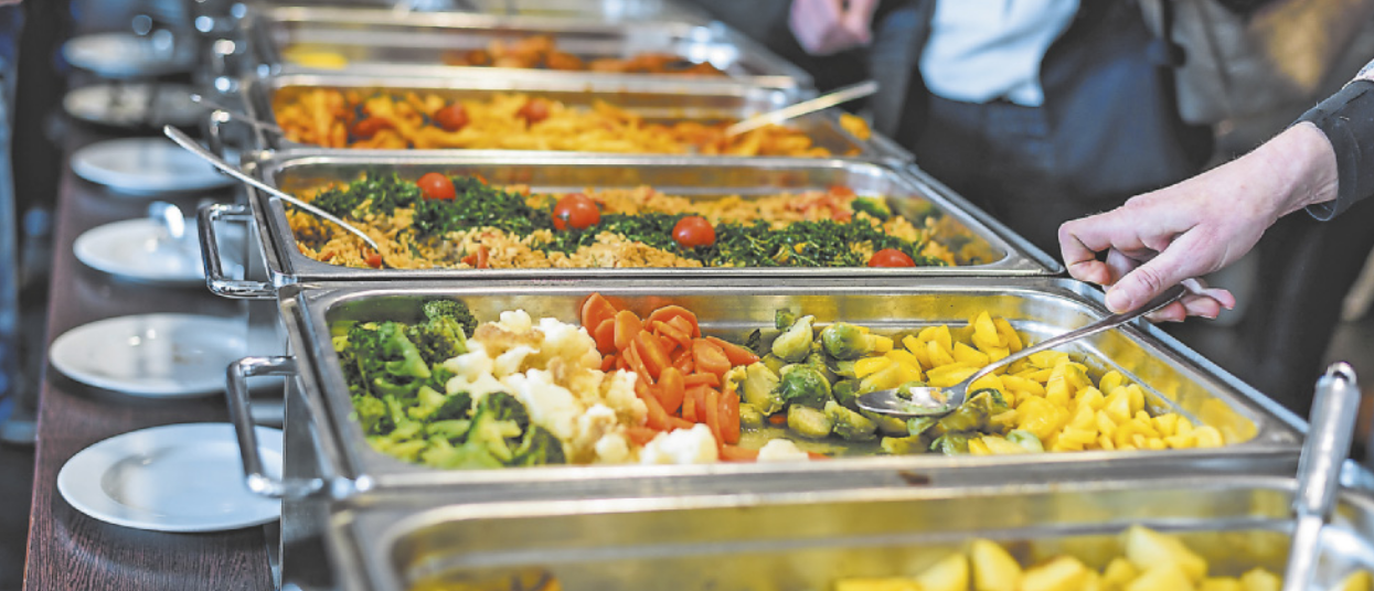 Fotografia de diversos alimentos dispostos em travessas de metal no estilo 'self-service'.