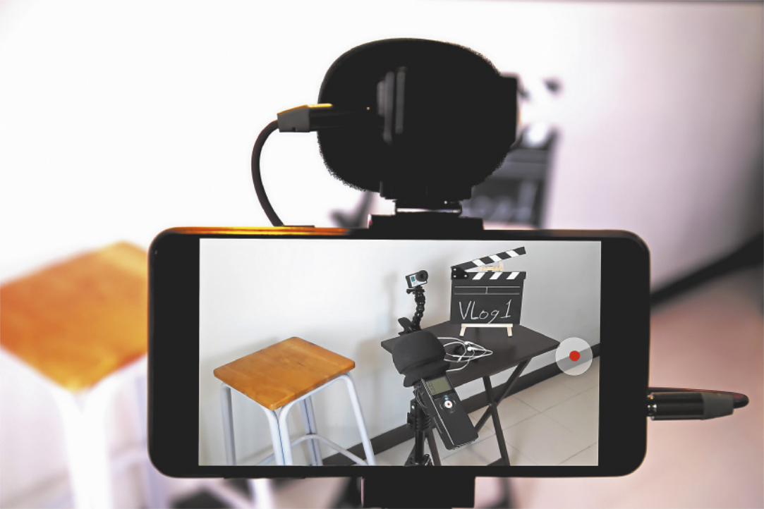 Fotografia da tela de um celular apoiado em um tripé, com a câmera apontada para um 'set' de gravação. O 'set' contém um banco, microfone, uma pequena câmera, fones de ouvido, e uma claquete com a mensagem: 'Vlog 1'.