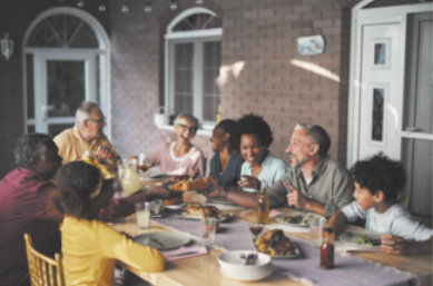Fotografia de uma família sentada à mesa para fazer uma refeição. A mesa está posta e as pessoas estão sorrindo e conversando entre si.