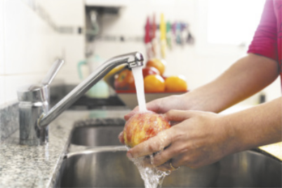 Fotografia de uma mulher lavando uma maçã na torneira da pia. Ao fundo, há outras frutas em uma fruteira.