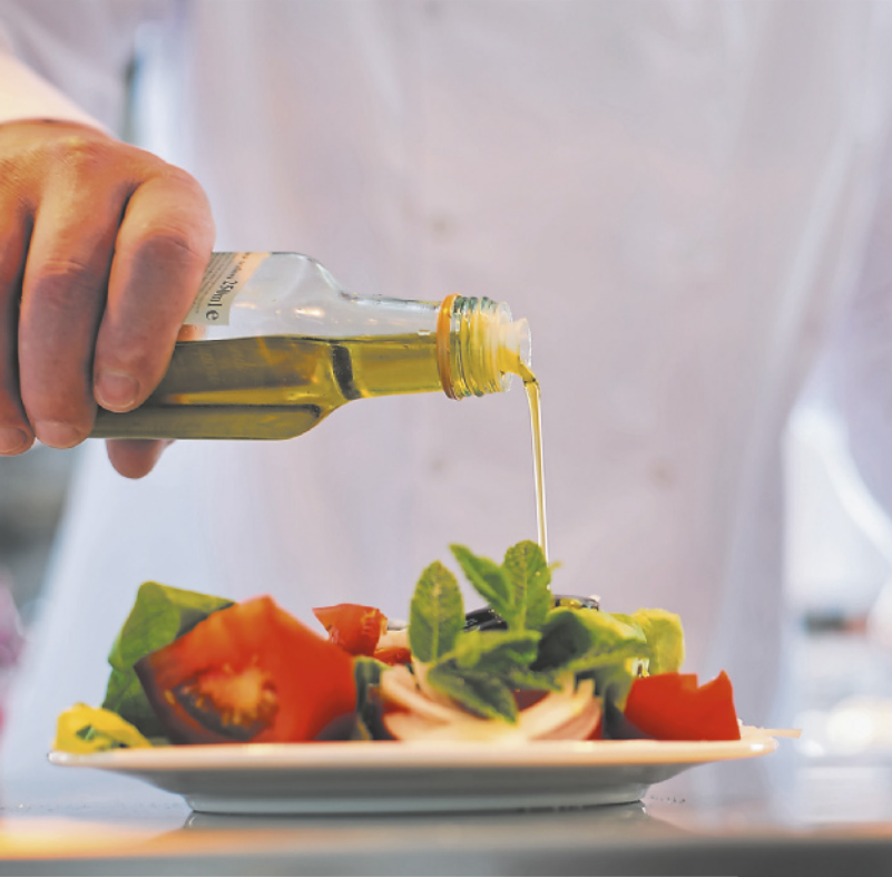Fotografia de uma pessoa despejando um fio de azeite em um prato de salada contendo folhas e tomate.