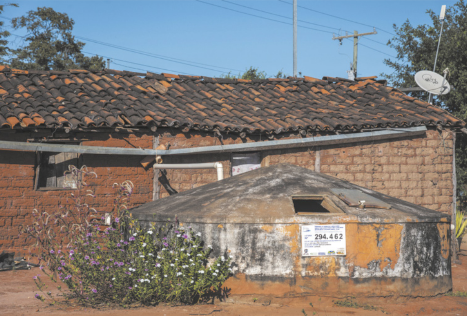 Fotografia de uma casa de tijolos com uma grande cisterna na frente. A cisterna é um recipiente esférico com uma tampa piramidal. Há algumas plantas na frente da casa.