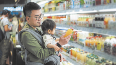 Fotografia de um homem segurando um bebê na frente do corpo, enquanto olha para uma garrafa de suco. Na frente do homem há prateleiras com uma diversidade de bebidas à venda.