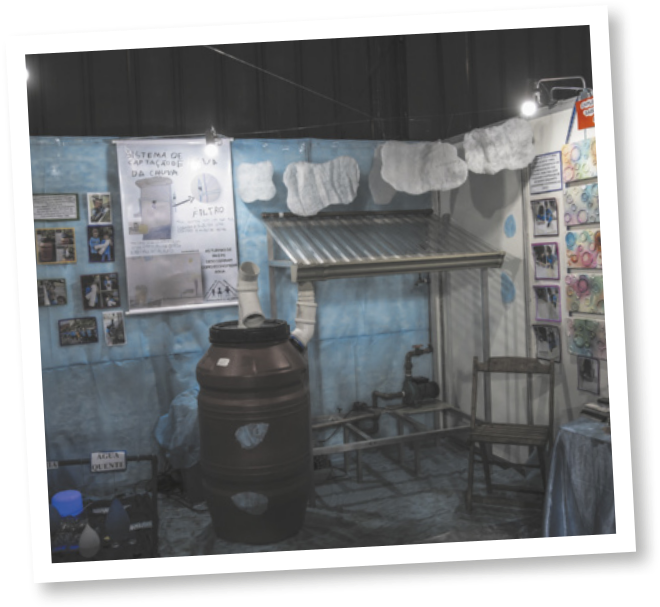 Fotografia de um pequeno telhado inclinado, com um cano que se conecta a um reservatório em formato de tambor. Há diversos papéis e fotos coladas nas paredes, e uma cadeira de madeira.