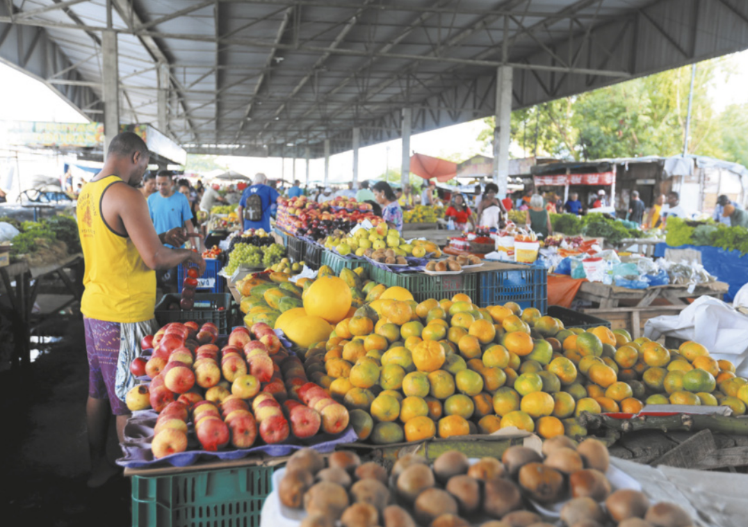 Fotografia de frutas e verduras em bancas de feira dispostas em um grande galpão. Há diversas pessoas observando os produtos.