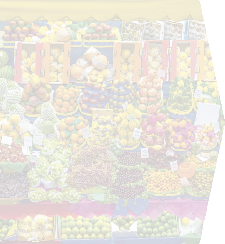 Fotografia de diversas frutas dispostas em uma banca. Há caixotes de papelão contendo frutas, e frutas empilhadas em pirâmides. Pequenas placas de papel identificam as frutas e os preços. A fotografia está com opacidade reduzida.