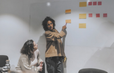 Fotografia de uma pessoa apresentando um storyboard, composto por vários post-its colados na parede. A mulher aponta para um dos post-its, enquanto outra mulher a escuta atentamente.