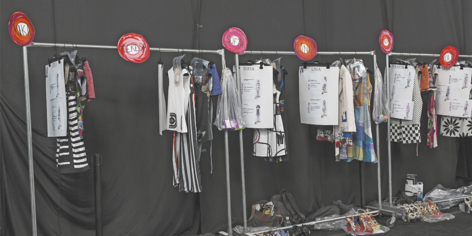 Fotografia de uma arara de roupas com peças separadas para cada pessoa. Para cada conjunto de roupas, há um papel contendo o nome e foto da pessoa que irá usá-lo.