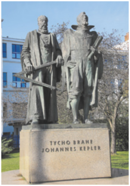 Fotografia de uma escultura retratando dois homens em pé. O homem da esquerda usa um traje longo e barba comprida. Ele segura um instrumento para medir ângulos e altitudes de corpos celestes. O homem à direita, está usando um traje mais formal e segurando um livro perto do corpo.