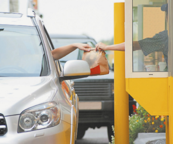 Fotografia da mão de uma pessoa entregando uma sacola de papel contendo 'fast-food' para uma pessoa em um carro. Atrás, há outro carro esperando sua vez.