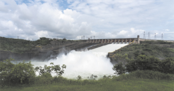 Fotografia de uma usina hidrelétrica. A barragem da hidrelétrica está com suas comportas abertas, liberando uma grande quantidade de água. Há vegetação no entorno e o céu está com nuvens.