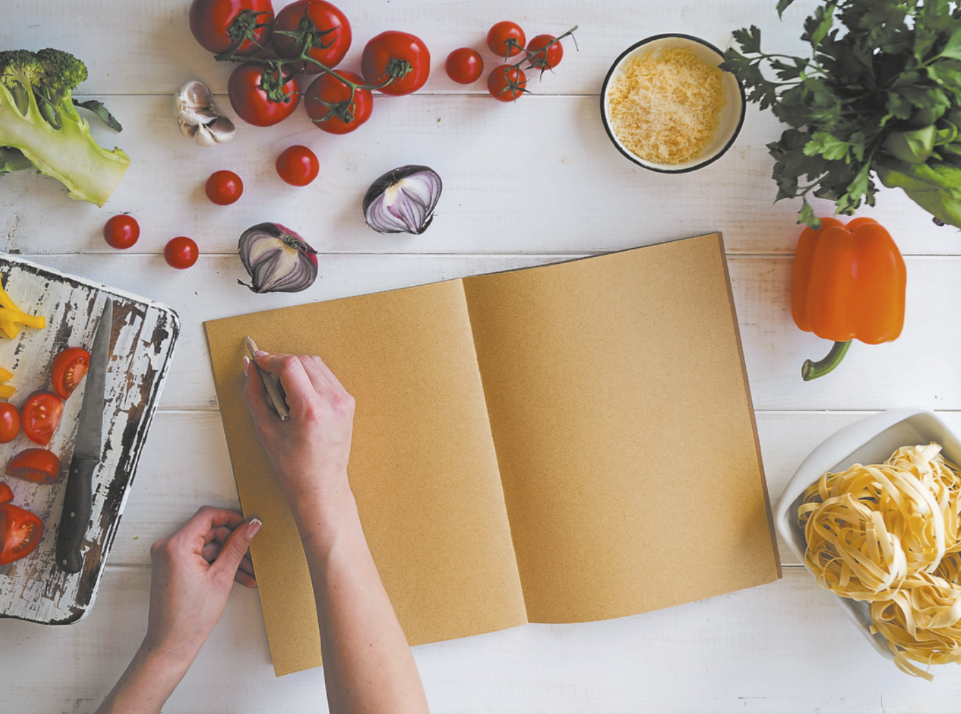 Fotografia da mão de uma pessoa anotando algo nas páginas de um caderno em branco. Ao redor do caderno, há diversos legumes em cima de uma mesa de madeira.