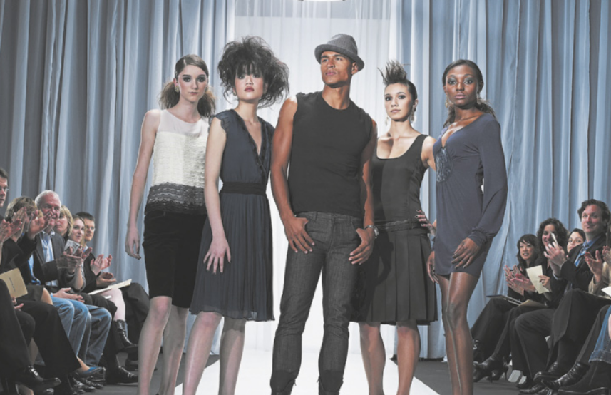 Fotografia de um grupo de modelos posando em uma passarela. Há plateia de ambos os lados, aplaudindo.