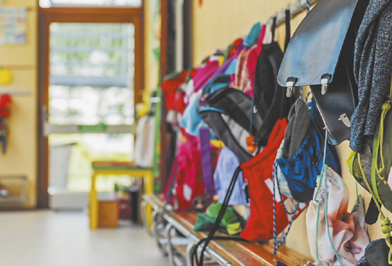 Fotografia de uma arara com várias mochilas penduradas. Abaixo das mochilas, há um banco de madeira, e ao fundo, uma porta que dá para a área externa.