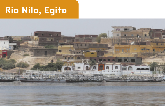 Fotografia 'Rio Nilo, Egito', representando um rio, cujas margens estão cobertas por barcos e construções baixas e compridas com muitas janelas.