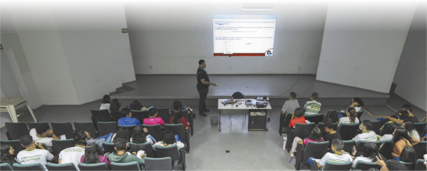 Fotografia de um homem fazendo uma apresentação de slides para uma sala de aula cheia. O apresentador aponta para algo nos slides, e a plateia está disposta em cadeiras em um espaço semelhante a um auditório. Há uma mesa na frente da projeção dos slides com vários aparelhos eletrônicos.