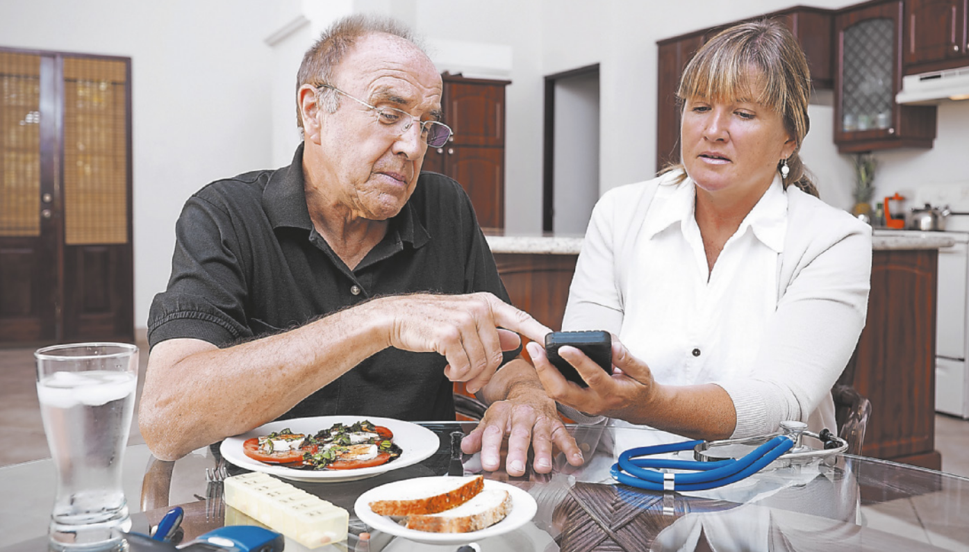 Fotografia de um homem idoso e uma mulher. À esquerda, o homem está sentado à mesa. Ele usa óculos e tem os cabelos grisalhos. Ele está apontando com o dedo indicador para um pequeno aparelho preto que a mulher está segurando. A mulher está sentada ao lado do homem, e tem cabelos loiros. Ela está segurando o aparelho e olhando para ele, com uma expressão focada. Em cima da mesa há pratos de comida, copos e um estetoscópio. Ao fundo, há armários, bancadas e portas.
