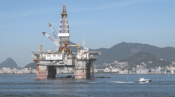 Fotografia de uma plataforma de petróleo. Ela é composta por uma estrutura elevada por pilastras acima do nível do mar. No meio da plataforma, há uma torre feita de metal. Ao fundo, há montanhas, edifícios e barcos.