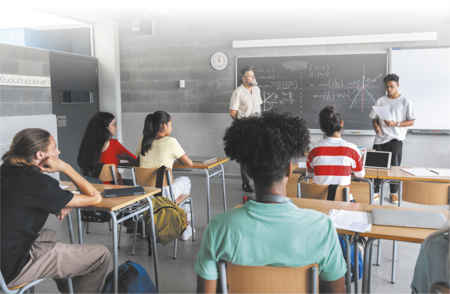 Fotografia de um jovem realizando uma apresentação para a classe. Ele segura uma folha de papel e a observa com expressão séria. À sua esquerda, um professor o escuta atentamente. À sua frente, os colegas estão sentados em carteiras escolares, acompanhando a apresentação com atenção. Sobre as carteiras, há notebooks, folhas de papel e canetas, enquanto mochilas estão penduradas nas cadeiras. Ao fundo, vê-se uma lousa com diversas equações e gráficos escritos, e, ao lado dela, um quadro branco.