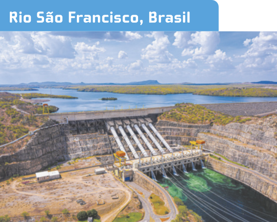 Fotografia aérea 'Rio São Francisco, Brasil', representando uma usina hidrelétrica. Atrás da barragem, a água está represada em um reservatório. A barragem principal é uma estrutura maciça de onde emergem várias tubulações inclinadas. Essas tubulações levam a água do reservatório para a casa de força, que está localizada na parte inferior da barragem. Na casa de força, há turbinas e geradores, e a água é despejada em um segundo reservatório. Nas laterais da barragem, há paredões rochosos e a vegetação parece típica de áreas mais secas. O céu está azul com muitas nuvens.