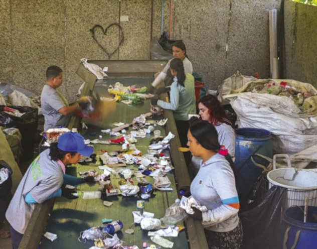 Fotografia de pessoas trabalhando em uma esteira com diversos produtos que serão reciclados. As pessoas estão usando luvas de proteção e selecionam alguns dos objetos que estão dispostos na esteira. De ambos os lados da esteira, há grandes tambores e sacos contendo os materiais recicláveis.