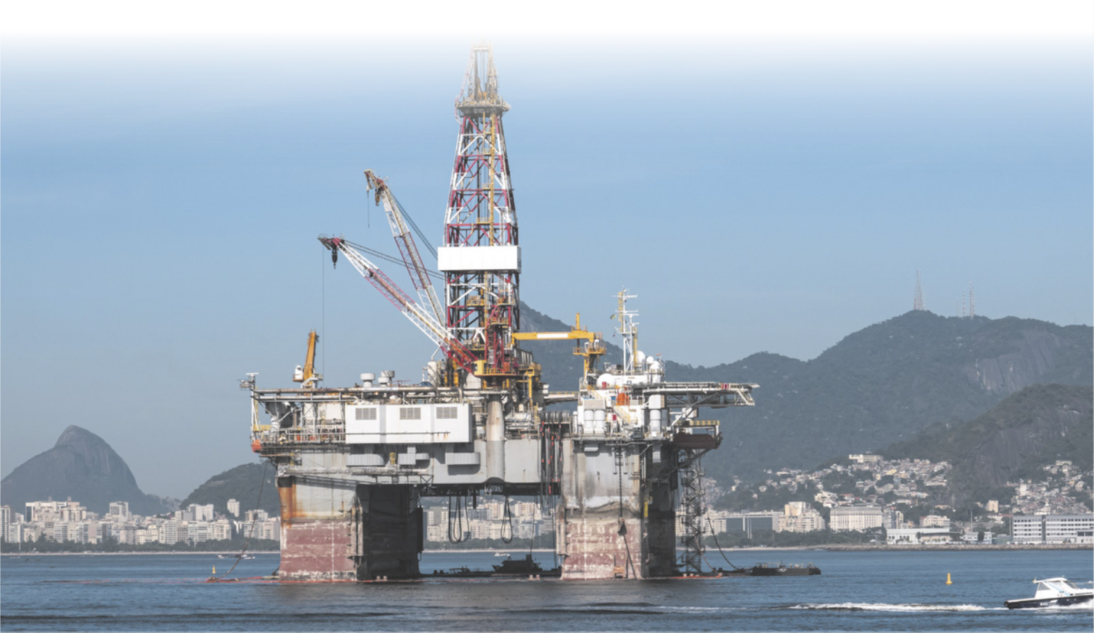 Fotografia de uma plataforma de petróleo. Ela é composta por uma estrutura elevada por pilastras acima do nível do mar. No meio da plataforma, há uma torre feita de metal. Ao fundo, há montanhas, uma cidade e barcos.