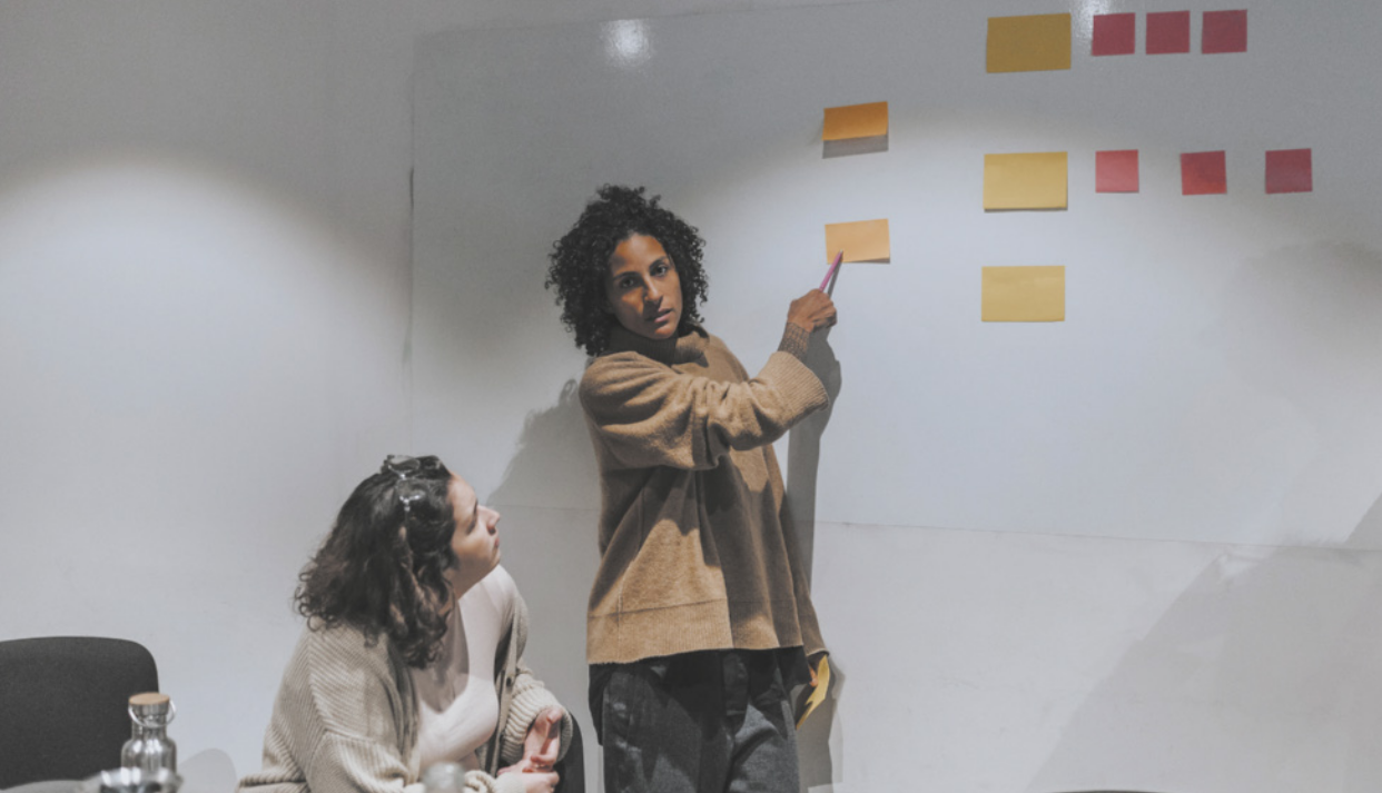 Fotografia de uma pessoa apresentando um storyboard, composto por vários post-its colados na parede. A mulher aponta para um dos post-its, enquanto outra mulher a escuta atentamente.