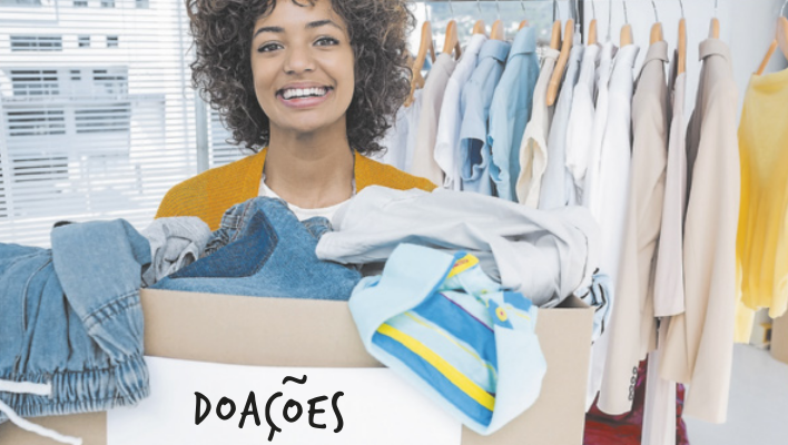 Fotografia de uma mulher segurando uma caixa de roupas, com uma etiqueta escrito: 'Doações'. Ao fundo, há uma arara com roupas penduradas.