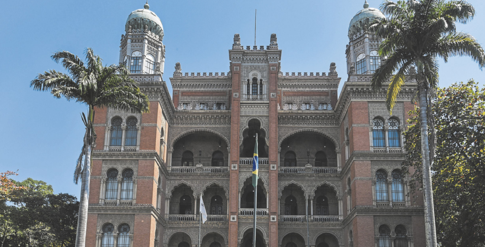 Fotografia de uma construção com duas torres e cúpulas . As múltiplas janelas e sacadas são ornamentadas com arcos, grades e detalhes decorativos. No centro da fachada, há um mastro com a bandeira do Brasil. Duas grandes palmeiras altas emolduram a fachada principal. Ao fundo, o céu está azul.