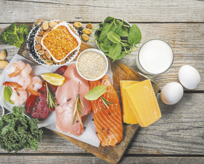 Fotografia de uma mesa de madeira contendo vários alimentos, como ovos, leite, queijos, carnes, folhas, legumes e grãos.