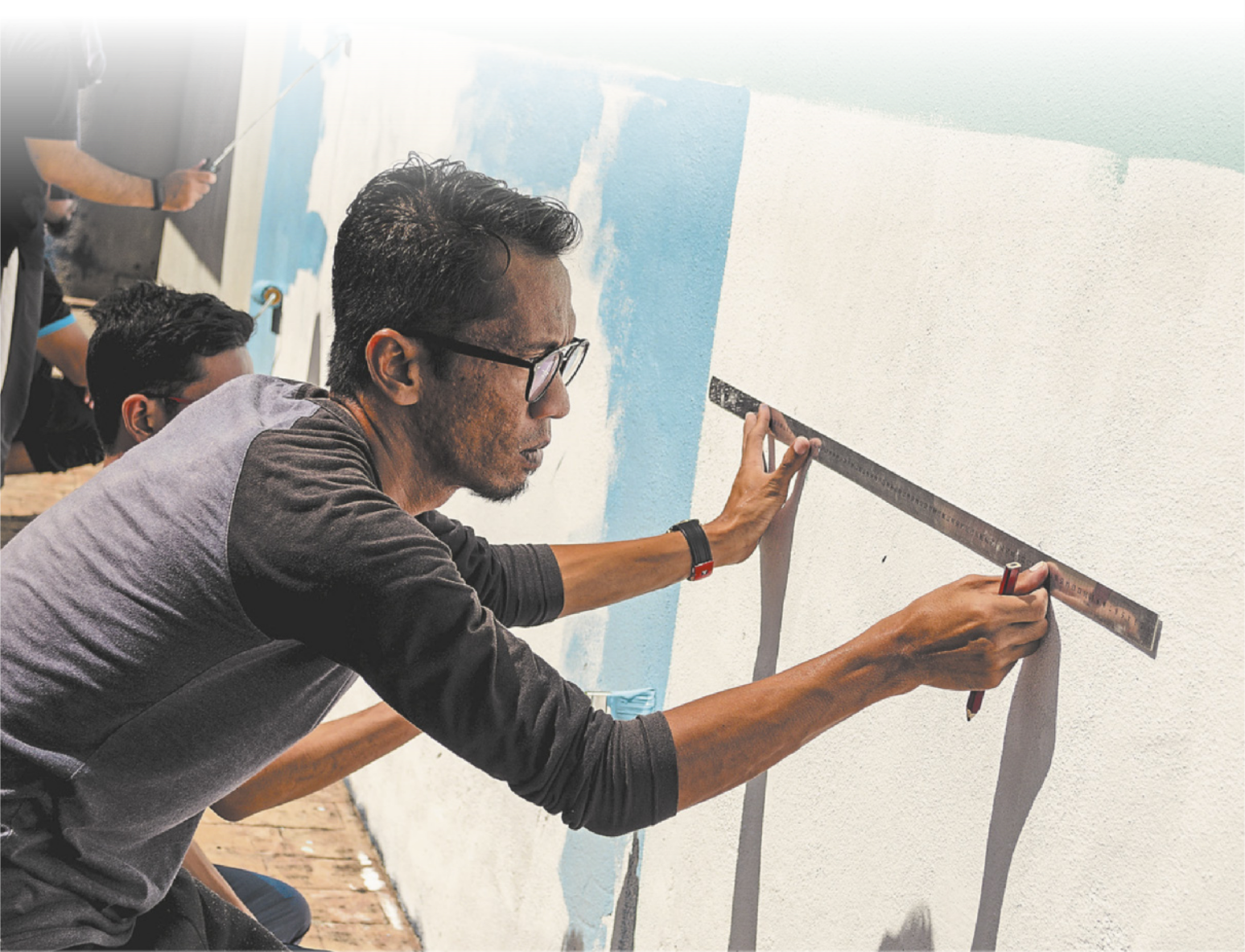 Fotografia de um homem usando uma régua para medir as dimensões em uma parede. A parede está parcialmente pintada, e há outras pessoas trabalhando na pintura.