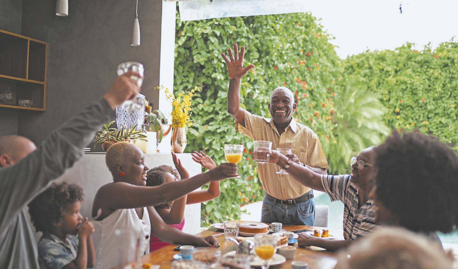 Fotografia de um grupo de pessoas brindando, reunidas na frente de uma mesa posta. Um dos homens está em pé com uma das mãos levantadas, enquanto brinda com a outra. Todos estão levantando seus copos em direção ao centro da mesa. Na mesa, há copos, pratos e um bolo. As pessoas estão com a expressão alegre. À esquerda, há uma estante na parede e luminárias no teto. À direita, há uma grande abertura que dá para um ambiente externo com muita vegetação.