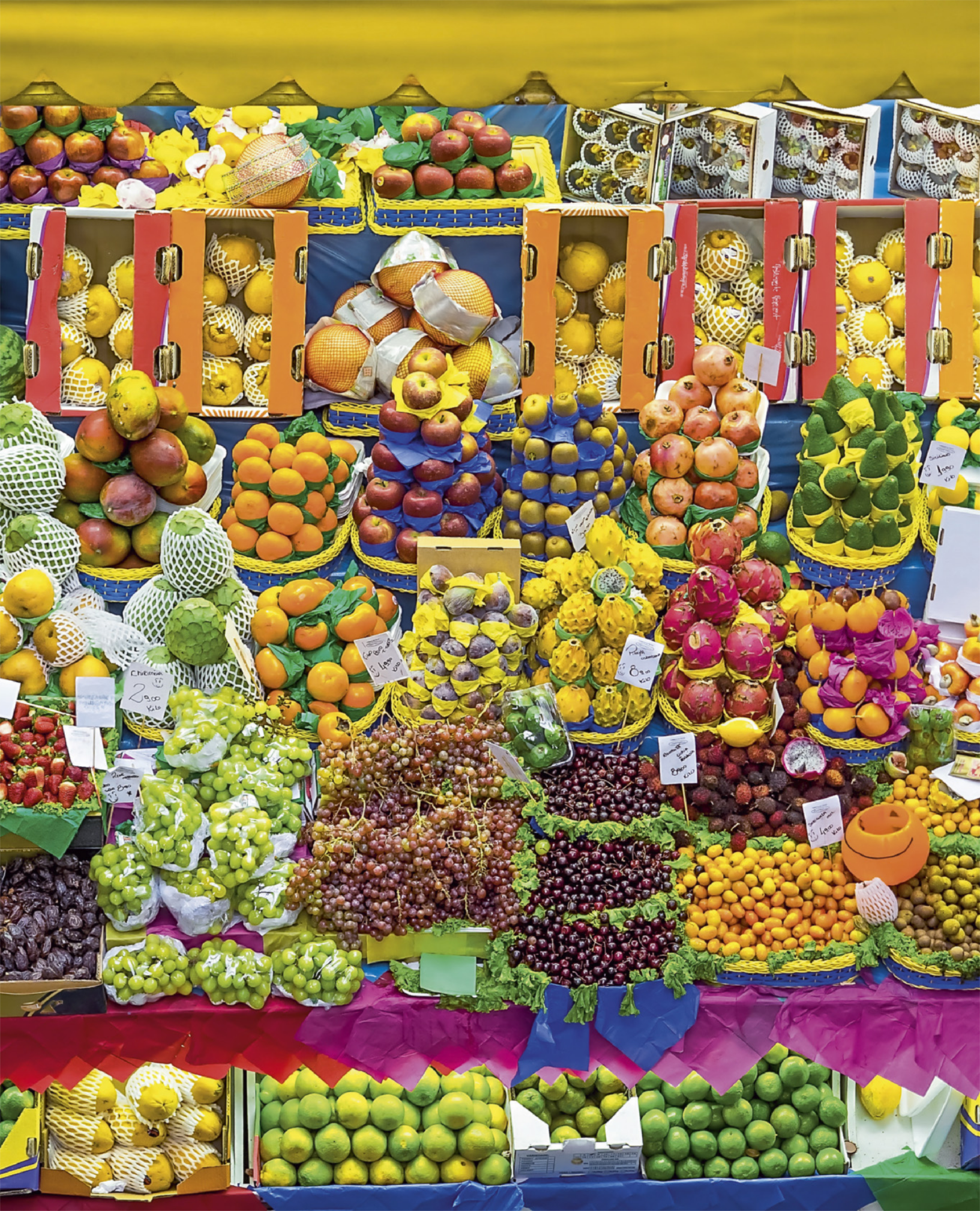 Fotografia de diversas frutas dispostas em uma banca. Há caixotes de papelão contendo frutas, e frutas empilhadas em pirâmides. Pequenas placas de papel identificam as frutas e os preços.