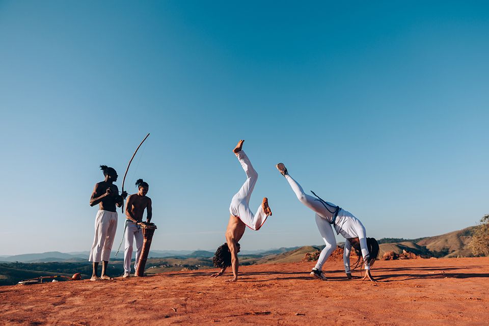 Fotografia. À direita, uma pessoa de blusa e calça branca está com o corpo inclinado para frente, as mãos no chão e uma perna para cima. Ao seu lado, uma pessoa de calça branca, de ponta cabeça com as mãos apoiadas no chão e as pernas para cima. À esquerda, dois homens de calça branca lado a lado, um deles toca berimbau e o outro tambor. O chão é de terra. Ao fundo, céu azul sem nuvens.