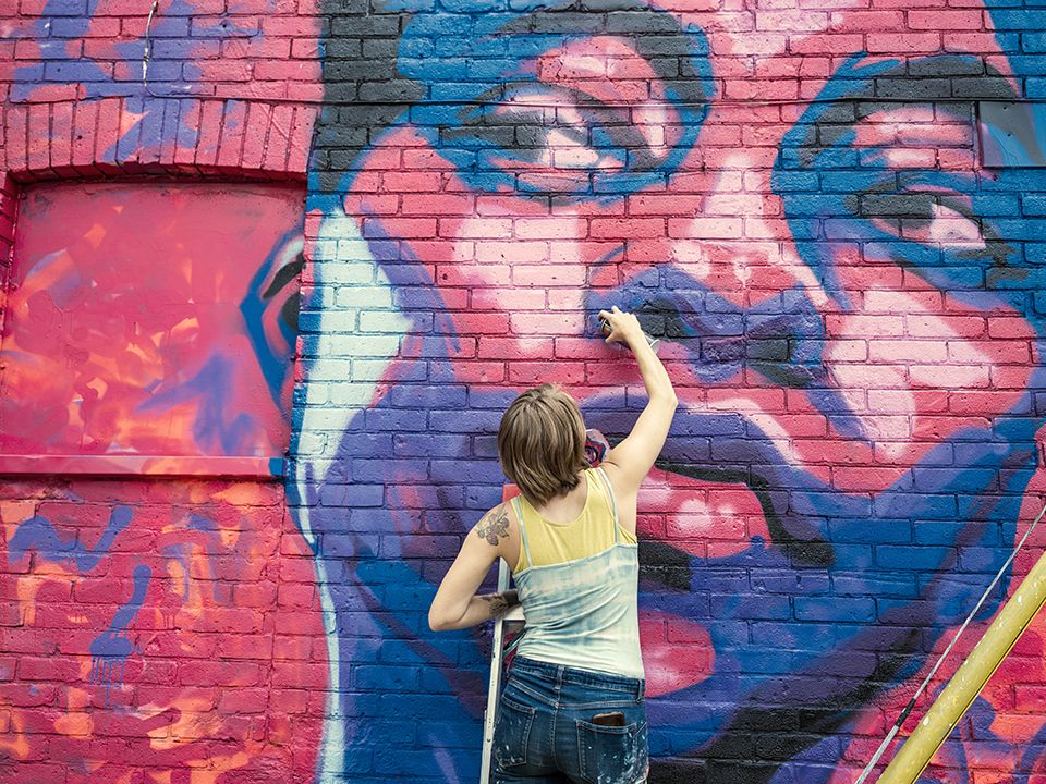 Fotografia. Uma mulher de cabelo castanho curto, vestindo blusa regata azul e amarela e calça jeans está de costas com uma mão estendida, segurando um spray de tinta próximo de um muro. No muro há o grafite do rosto de uma pessoa em tons de roxo e rosa.