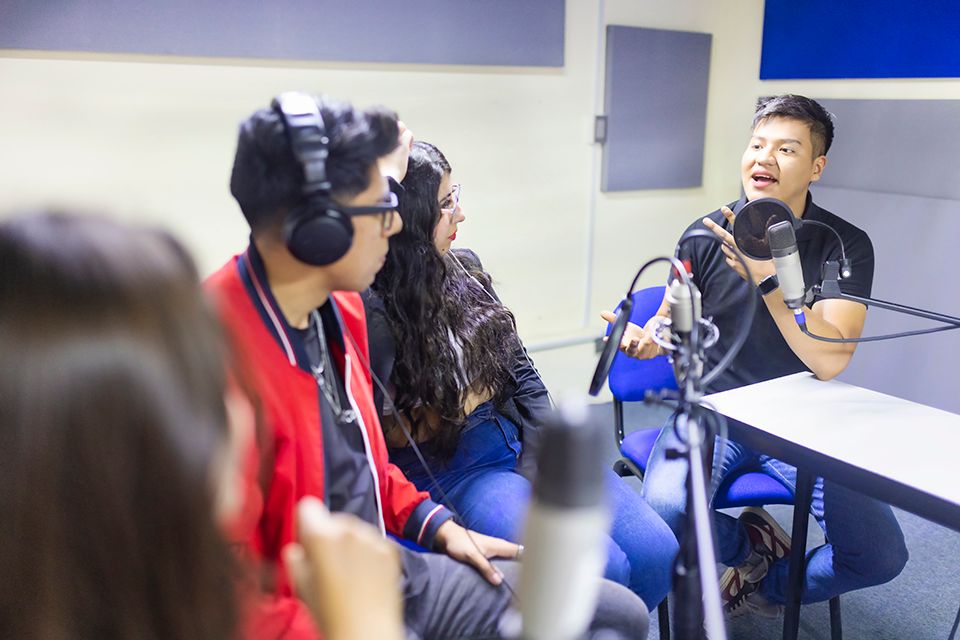 Fotografia. À direita, um homem de cabelo preto e curto vestindo camiseta preta e calça jeans está sentado ao lado de uma mesa. Na sua frente há um microfone. À esquerda, uma menina de óculos, cabelo castanho ondulado, blusa preta e calça jeans e um homem de cabelo castanho e curto, usando óculos, fones de ouvido, casaco vermelho e calça preta estão olhando para o primeiro homem. No canto esquerdo, há uma pessoa de cabelo castanho de costas.
