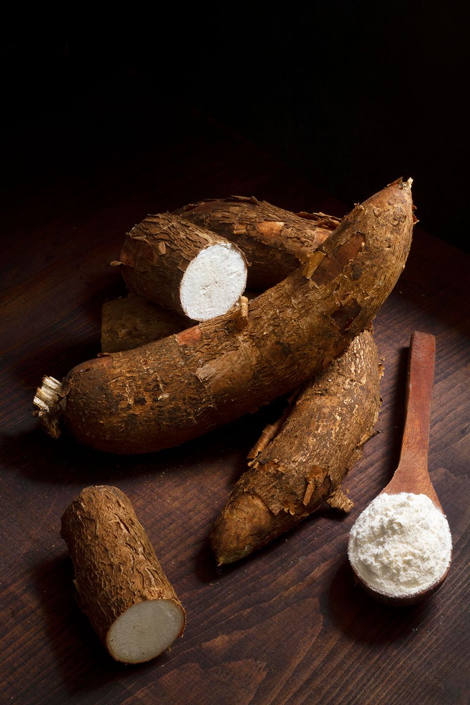 Fotografia. Mandiocas, raiz de formato cilíndrico com camada externa marrom e miolo amarelo. À direita, uma colher com farinha de grãos amarelos.