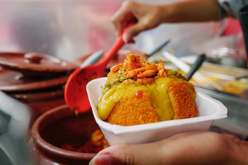 Fotografia. Destaque para as mãos de uma pessoa, com uma mão ela segura uma colher em cima de uma panela, com a outra mão ela segura uma bandeja branca com um acarajé com vatapá de camarão dentro.