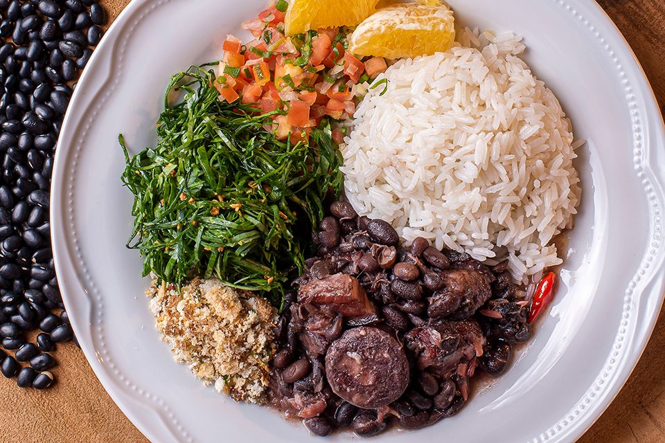 Fotografia. Vista de cima de um prato com arroz, feijoada, farofa, couve, vinagrete e gomos de laranja.