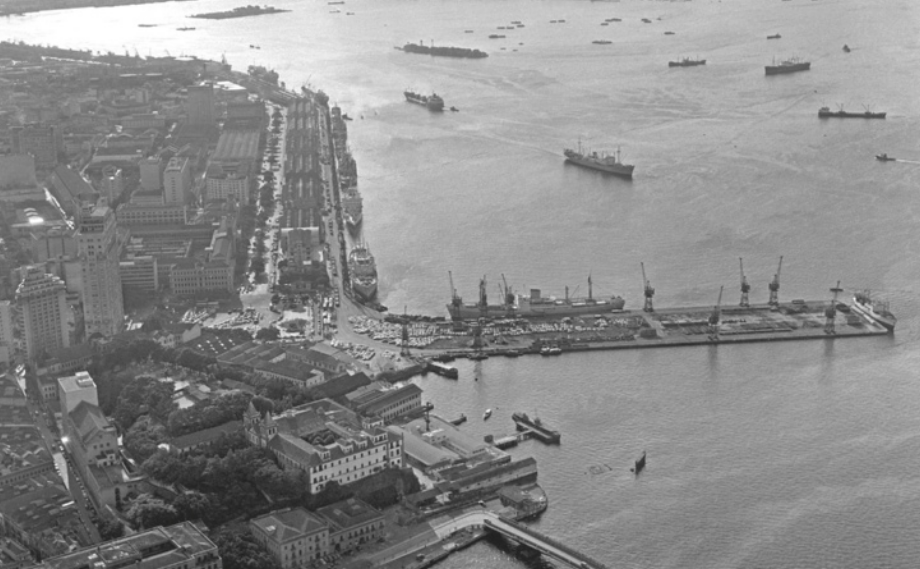 Fotografia aérea em preto e branco de uma região portuária, com vista para a cidade e o mar. À esquerda, observa-se parte da área urbana, com edifícios e ruas organizadas. Próximo à margem, há galpões, guindastes e navios atracados. No mar, encontram-se diversas embarcações de grande porte, tanto ancoradas quanto em alto-mar.