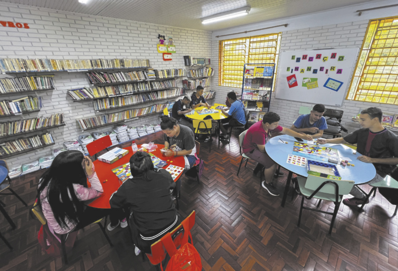 Fotografia de jovens sentados ao redor de três mesas redondas em uma biblioteca, jogando jogos de tabuleiro.