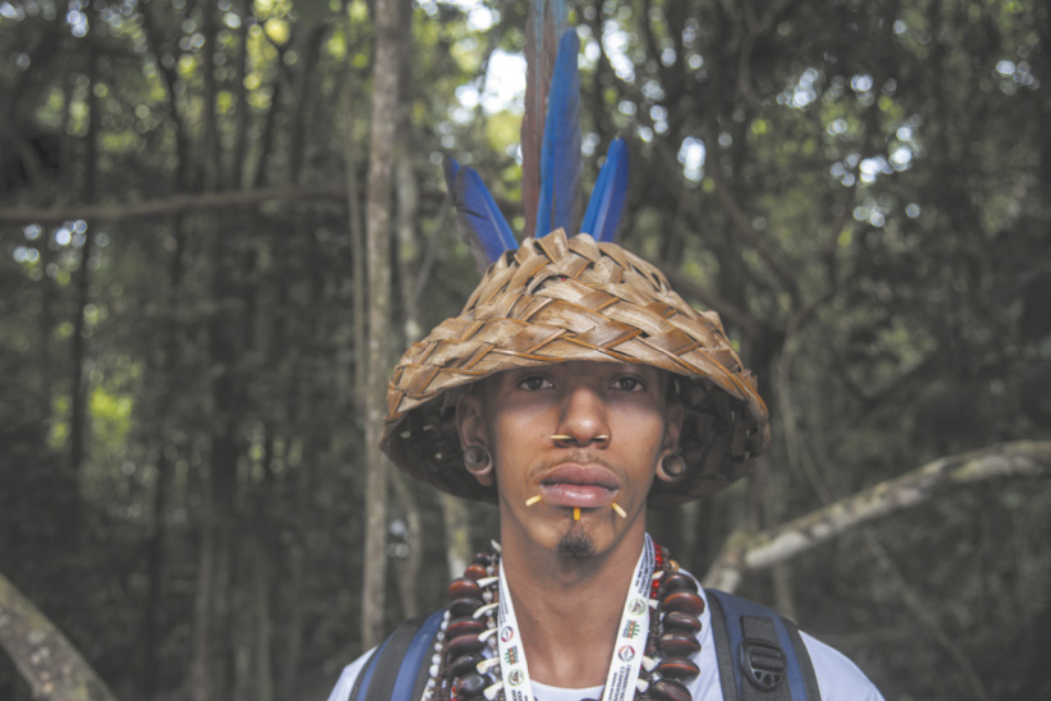 Fotografia de um jovem indígena em uma área de mata fechada, usando chapéu de palha com três penas, colar de sementes e adornos de madeira perfurando o nariz e o lábio inferior.