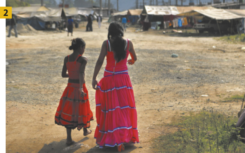 Fotografia 2 mostra uma mulher e uma menina vestindo saias longas e coloridas, além de blusinhas, caminhando por um chão de terra batida em direção a um acampamento com tendas e roupas penduradas em varais.