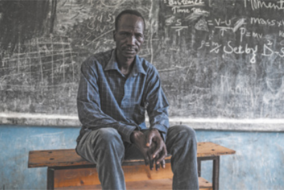 Fotografia de um homem negro com expressão séria, vestindo camisa de manga comprida e calça, sentado sobre uma mesa, tendo ao fundo uma lousa com marcas de giz.