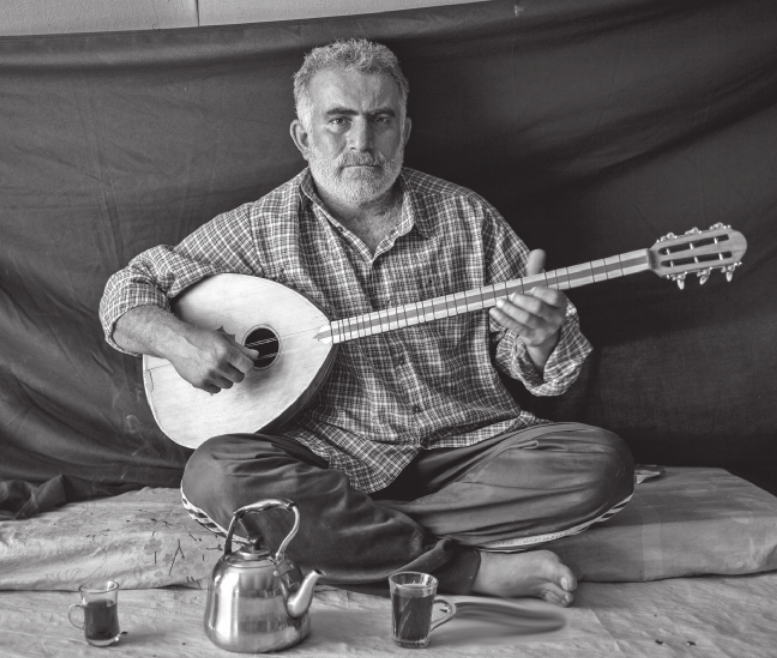 Fotografia em preto e branco de homem sentado no chão com as pernas cruzadas, tocando um instrumento musical de cordas semelhante a um alaúde. À sua frente, sobre o chão, há uma chaleira metálica e dois copos pequenos com bebida. O ambiente é simples, com um tecido estendido como fundo.