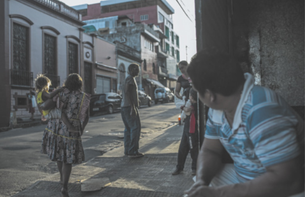 Fotografia de uma rua em área urbana. Uma mulher caminha carregando uma criança no colo. Outras pessoas estão paradas na calçada e no meio da rua, conversando. Um homem, sentado em frente a um estabelecimento, observa a cena.