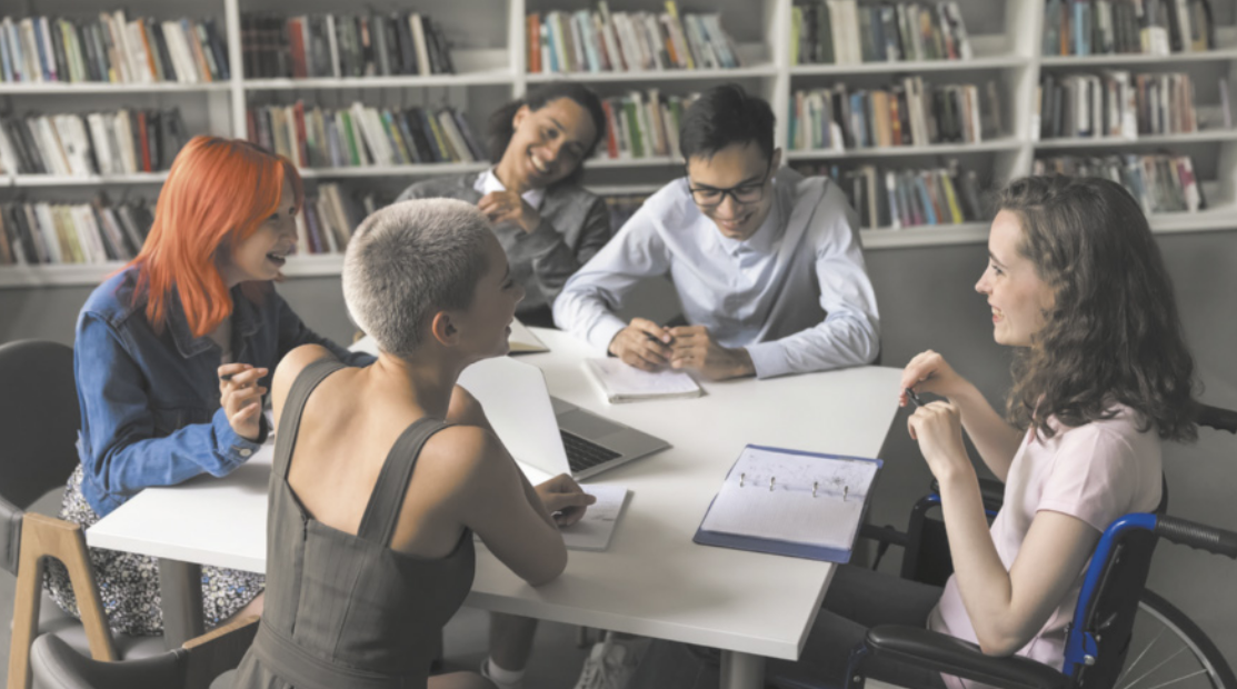 Fotografia de um grupo de estudantes sentados ao redor de uma mesa em uma biblioteca, conversando, sorrindo e escrevendo em seus cadernos.