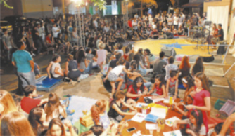 Fotografia de um grande número de pessoas reunidas em um espaço público ao ar livre. Há adultos, jovens e crianças sentados no chão, em mesas e ao redor de um pequeno palco, onde algumas pessoas tocam instrumentos. Próximas ao palco, algumas crianças desenham e pintam sentadas no chão.